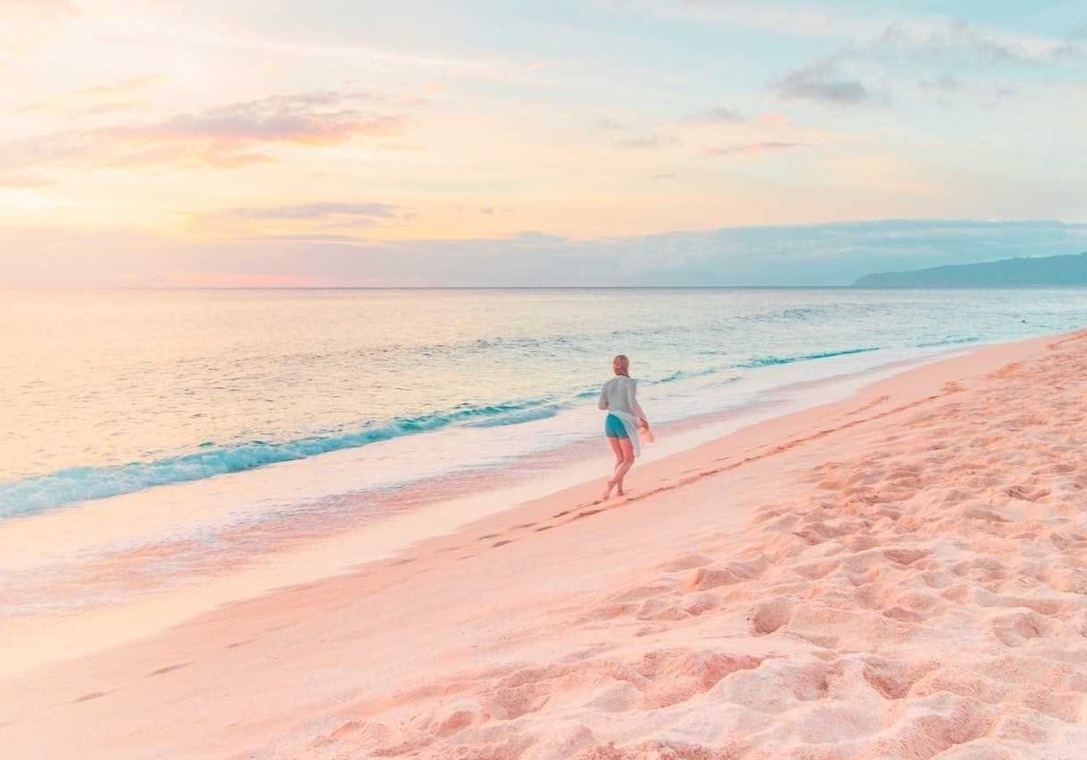 Woman walking along beach with pink sand against sunset.